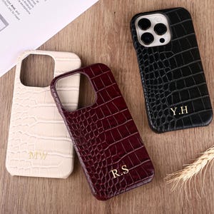 May include: Three phone cases with a crocodile skin texture are displayed on a wooden surface. The cases are in cream, burgundy, and black. Each case has gold-colored initials: "MW", "R.S", and "Y.H".