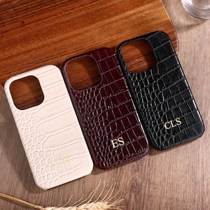 May include: Three phone cases with a crocodile skin texture. One is off-white, one is burgundy, and one is black. The burgundy case has the initials "ES" in gold, and the black case has "CLS" in gold. The cases are on a wooden surface.