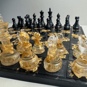 Peut inclure: Un jeu d'échecs noir et or avec des pièces en résine transparente. Les pièces d'échecs sont remplies de feuilles d'or et reposent sur un échiquier noir. Les pièces sont noires et dorées.