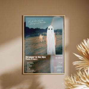 Puede incluir: Un póster enmarcado con la portada del álbum "Stranger in the Alps" de Phoebe Bridgers. La obra de arte muestra a una persona con un disfraz de fantasma en un campo con un perro. El póster incluye la lista de canciones y la fecha de lanzamiento.
