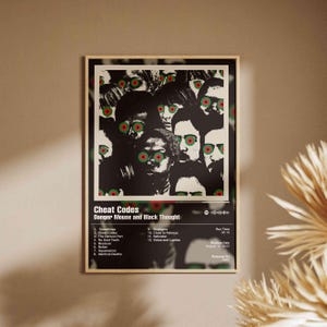 Puede incluir: Un póster enmarcado con la portada del álbum "Cheat Codes" de Danger Mouse y Black Thought. La obra de arte es un collage en blanco y negro de caras con círculos rojos y verdes para los ojos. El título del álbum y la lista de canciones se muestran debajo.