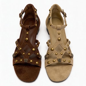 Op de afbeelding: Een paar platte sandalen in donkerbruin en beige, met een T-bandontwerp en een ruitpatroon aan de voorzijde. De bandjes zijn versierd met ronde metalen studs en kleine textuuraccenten, en worden vastgemaakt met een verstelbaar enkelbandje met gesp.