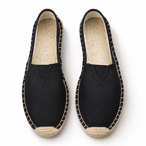 K&ouml;nnte beinhalten: Ein Paar schwarze Slipper-Espadrilles mit strukturiertem Stoffobermaterial und einem V-f&ouml;rmigen elastischen Einsatz am Rist. Die Schuhe haben eine Sohle aus naturfarbenem Juteseil mit sichtbaren Steppn&auml;hten am Rand und einer verst&auml;rkten Zehenkappe.