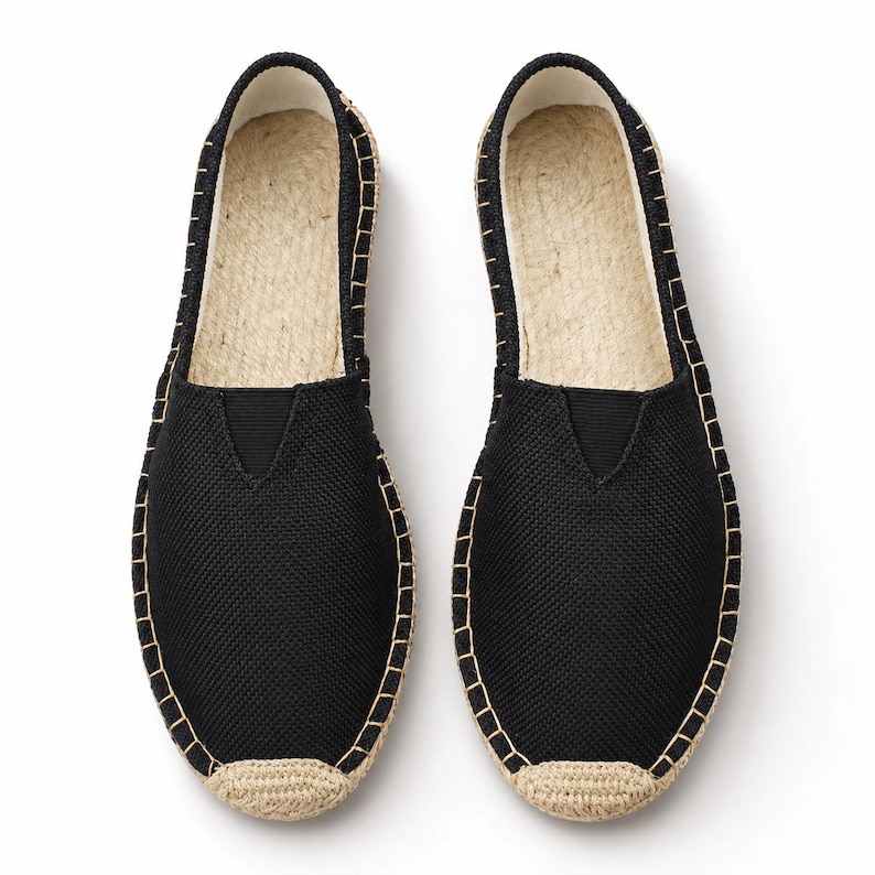 K&ouml;nnte beinhalten: Ein Paar schwarze Slipper-Espadrilles mit strukturiertem Stoffobermaterial und einem V-f&ouml;rmigen elastischen Einsatz am Rist. Die Schuhe haben eine Sohle aus naturfarbenem Juteseil mit sichtbaren Steppn&auml;hten am Rand und einer verst&auml;rkten Zehenkappe.