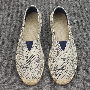 K&ouml;nnte beinhalten: Ein Paar Espadrilles zum Hineinschl&uuml;pfen mit einem cremefarbenen Obermaterial aus Segeltuch und einem schwarzen Gekritzel-Muster. Die Schuhe haben einen dunkelblauen, dreieckigen elastischen Einsatz am Spann und eine Sohle aus geflochtenem Juteseil.