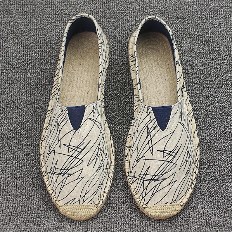 K&ouml;nnte beinhalten: Ein Paar Espadrilles zum Hineinschl&uuml;pfen mit einem cremefarbenen Obermaterial aus Segeltuch und einem schwarzen Gekritzel-Muster. Die Schuhe haben einen dunkelblauen, dreieckigen elastischen Einsatz am Spann und eine Sohle aus geflochtenem Juteseil.