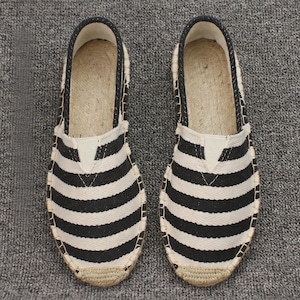 K&ouml;nnte beinhalten: Ein Paar Slipper-Espadrilles mit einem Obermaterial aus schwarz-wei&szlig; gestreiftem Canvas, einer runden Zehenpartie und einer Sohle aus geflochtenem Juteseil. Die Schuhe haben einen schwarzen Stoffbesatz am Einstieg und eine verst&auml;rkte Jutekappe.