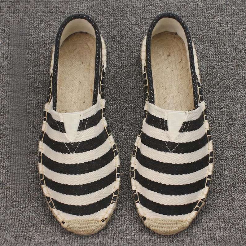 K&ouml;nnte beinhalten: Ein Paar Slipper-Espadrilles mit einem Obermaterial aus schwarz-wei&szlig; gestreiftem Canvas, einer runden Zehenpartie und einer Sohle aus geflochtenem Juteseil. Die Schuhe haben einen schwarzen Stoffbesatz am Einstieg und eine verst&auml;rkte Jutekappe.