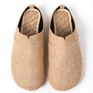 May include: A pair of slip-on shoes featuring a tan, textured burlap-like exterior fabric. The interior footbed is lined with a woven, light-colored natural fiber material in a herringbone pattern, with a small pull tab at the heel.