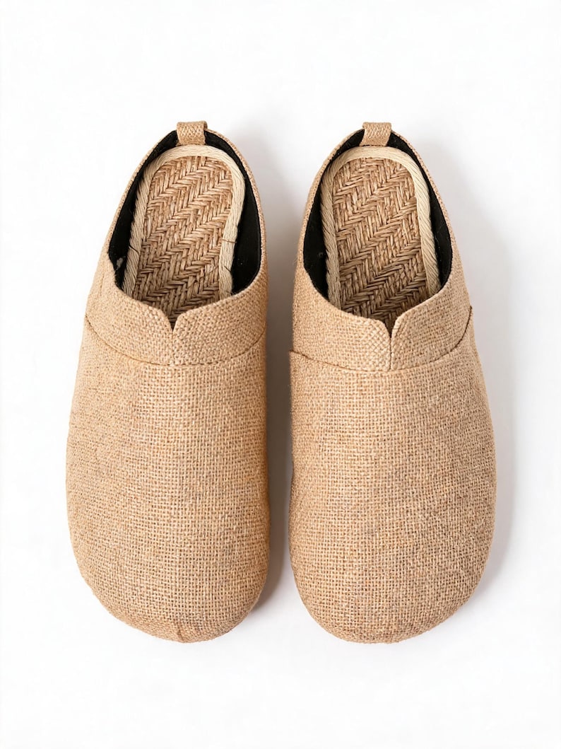 May include: A pair of slip-on shoes featuring a tan, textured burlap-like exterior fabric. The interior footbed is lined with a woven, light-colored natural fiber material in a herringbone pattern, with a small pull tab at the heel.