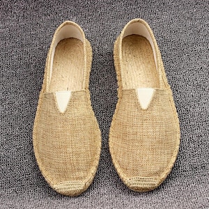 K&ouml;nnte beinhalten: Ein Paar beigefarbene Slipper mit einem Obermaterial aus strukturiertem Webstoff, einer abgerundeten Schuhspitze und einer verst&auml;rkten, mit Jute umwickelten Sohle. Jeder Schuh verf&uuml;gt &uuml;ber einen kleinen wei&szlig;en elastischen Einsatz am Rist.