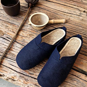 May include: A pair of dark navy blue slip-on shoes with a textured fabric exterior and a light beige woven straw-like insole. The shoes feature a rounded toe design and a low-cut opening, showcasing a simple construction without laces or straps.