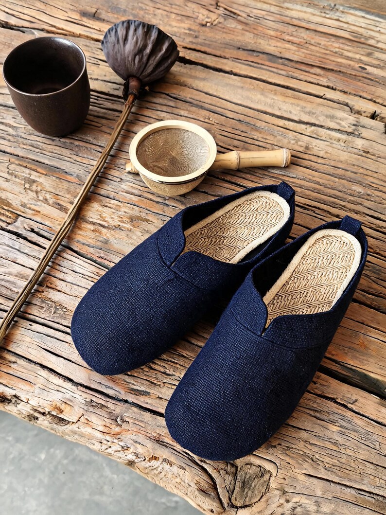 May include: A pair of dark navy blue slip-on shoes with a textured fabric exterior and a light beige woven straw-like insole. The shoes feature a rounded toe design and a low-cut opening, showcasing a simple construction without laces or straps.