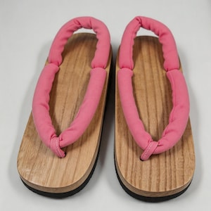 May include: A pair of geta sandals featuring light brown wooden platforms with a visible grain pattern, black rubber outsoles, and thick, padded pink fabric thong straps that loop around the heel for a secure fit on the foot.