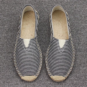 K&ouml;nnte beinhalten: Ein Paar Slipper mit einem Obermaterial aus schwarz-wei&szlig; gestreiftem Stoff, einer beigen Sohlenkante in Jute-Optik und einem hellen elastischen Einsatz am Rist. Das Innenfutter der Sohle besteht aus einem naturfarbenen, gewebten Material.