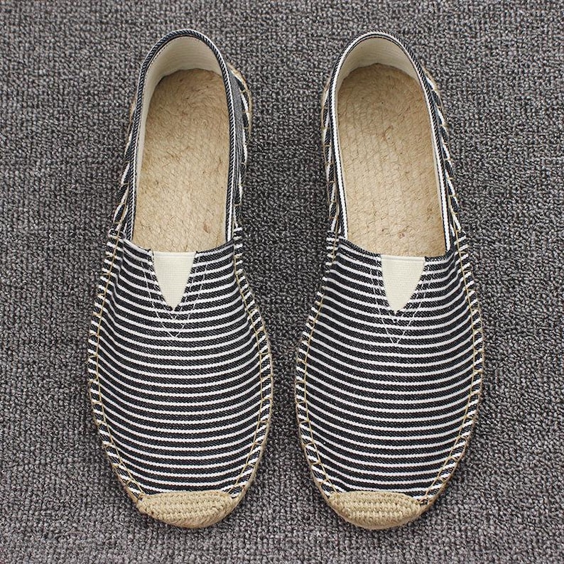 K&ouml;nnte beinhalten: Ein Paar Slipper mit einem Obermaterial aus schwarz-wei&szlig; gestreiftem Stoff, einer beigen Sohlenkante in Jute-Optik und einem hellen elastischen Einsatz am Rist. Das Innenfutter der Sohle besteht aus einem naturfarbenen, gewebten Material.