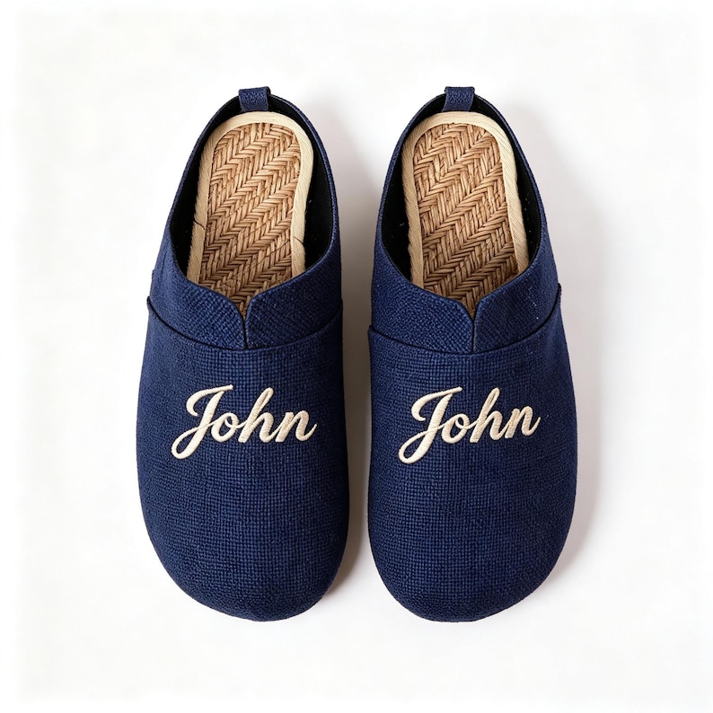 May include: A pair of navy blue slip-on slippers featuring the name John embroidered in white script on the top of each shoe. The interior footbed has a tan herringbone woven texture, and the exterior is made of a solid navy blue fabric.