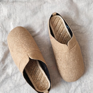 May include: A pair of slip-on shoes featuring a textured tan fabric upper and a woven jute footbed. The shoes have a rounded toe design, a black inner lining, and a small pull tab at the heel, showcasing a natural, neutral color palette.