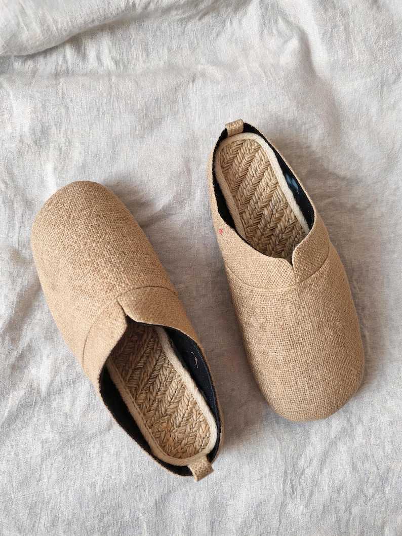 May include: A pair of slip-on shoes featuring a textured tan fabric upper and a woven jute footbed. The shoes have a rounded toe design, a black inner lining, and a small pull tab at the heel, showcasing a natural, neutral color palette.