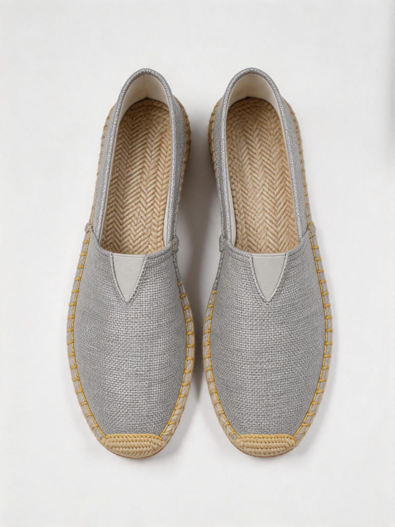 K&ouml;nnte beinhalten: Ein Paar silberne Slipper mit strukturiertem Stoffobermaterial, einem V-f&ouml;rmigen elastischen Einsatz und einer Zwischensohle aus geflochtener Jute mit gelben N&auml;hten am Rand. Das Innere ist mit einer Fischgr&auml;t-Innensohle ausgestattet.