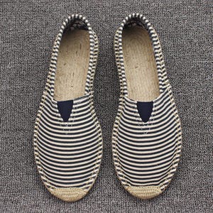 K&ouml;nnte beinhalten: Ein Paar Slipper-Espadrilles mit einem Obermaterial aus marineblau-wei&szlig; gestreiftem Canvas, einer abgerundeten Schuhspitze, einem kleinen, einfarbig marineblauen, dreieckigen elastischen Einsatz am Rist und einer Sohle aus geflochtenem Juteseil.