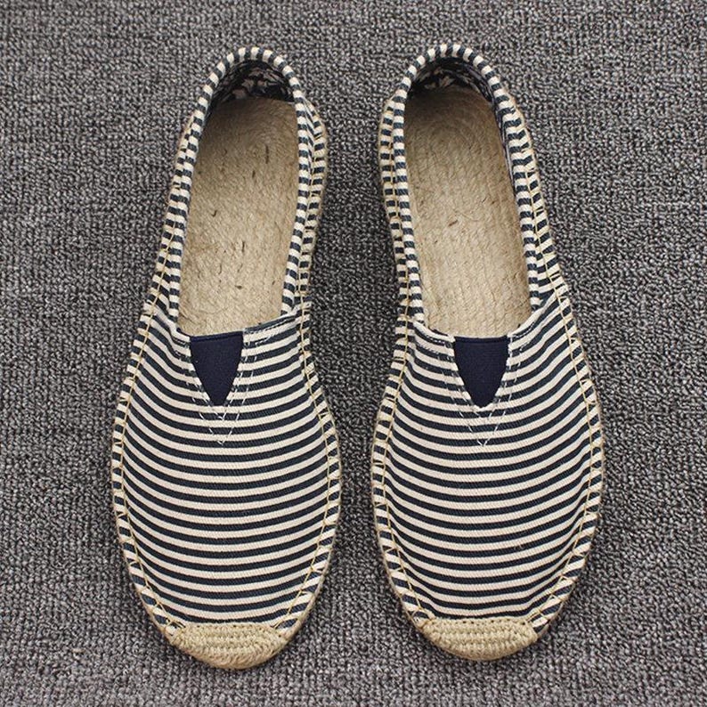 K&ouml;nnte beinhalten: Ein Paar Slipper-Espadrilles mit einem Obermaterial aus marineblau-wei&szlig; gestreiftem Canvas, einer abgerundeten Schuhspitze, einem kleinen, einfarbig marineblauen, dreieckigen elastischen Einsatz am Rist und einer Sohle aus geflochtenem Juteseil.