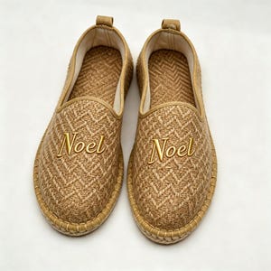 May include: A pair of tan slip-on shoes featuring a woven herringbone texture and the word Noel embroidered in gold thread on the top of each shoe. The shoes have a rounded toe, a light-colored interior lining, and a stitched trim along the sole.