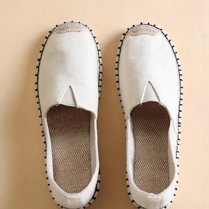 Può includere: Un paio di scarpe slip-on color bianco sporco con punta arrotondata, apertura a V sul collo del piede e fodera interna color marrone chiaro testurizzata. I bordi esterni delle suole presentano un punto a festone nero e punta intrecciata.