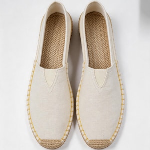 K&ouml;nnte beinhalten: Ein Paar cremewei&szlig;e Slipper aus Segeltuch mit einer Sohle aus geflochtenem Jute und einem strukturierten Fischgr&auml;tenmuster auf der Innensohle. Die Au&szlig;enkanten sind mit sichtbaren gelben N&auml;hten versehen, die das Obermaterial befestigen.