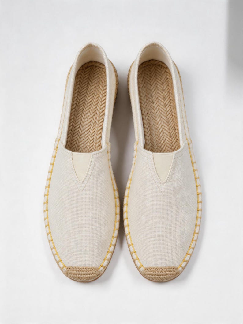 K&ouml;nnte beinhalten: Ein Paar cremewei&szlig;e Slipper aus Segeltuch mit einer Sohle aus geflochtenem Jute und einem strukturierten Fischgr&auml;tenmuster auf der Innensohle. Die Au&szlig;enkanten sind mit sichtbaren gelben N&auml;hten versehen, die das Obermaterial befestigen.