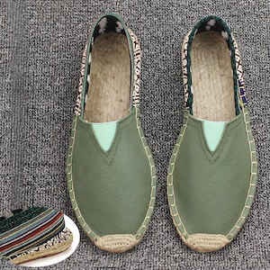 K&ouml;nnte beinhalten: Ein Paar olivgr&uuml;ne Slipper mit glatter Vorderseite, einem hellgr&uuml;nen V-f&ouml;rmigen Einsatz und einer Sohlenkante aus geflochtener Jute. Der Fersenbereich zeigt einen gemusterten Stoff mit wei&szlig;en, schwarzen und kastanienbraunen geometrischen Designs.