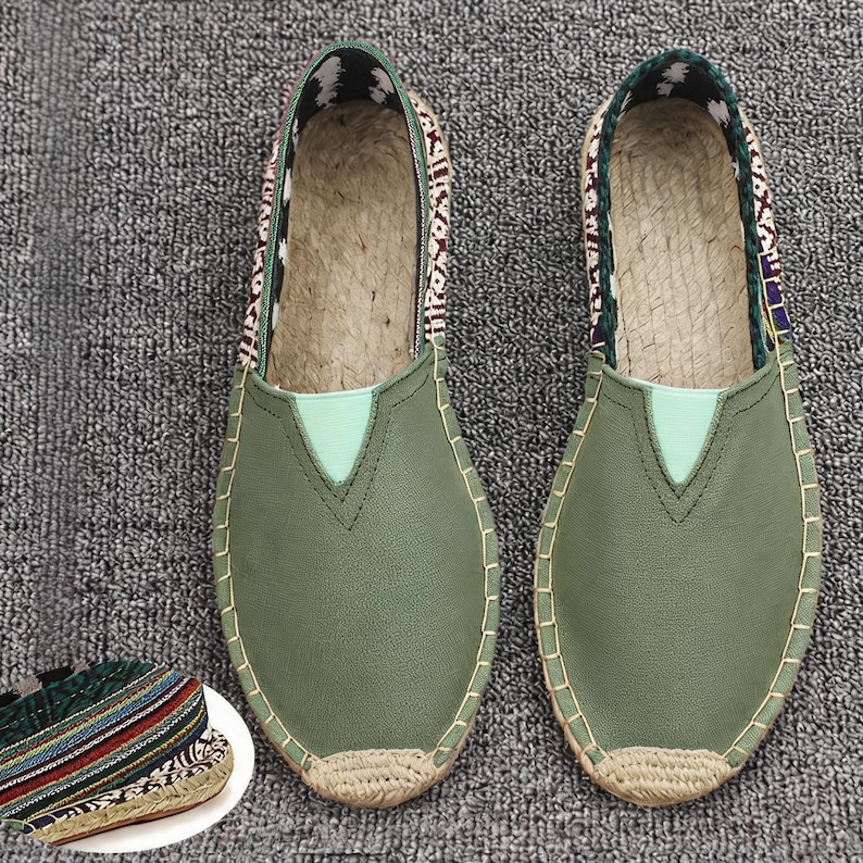 K&ouml;nnte beinhalten: Ein Paar olivgr&uuml;ne Slipper mit glatter Vorderseite, einem hellgr&uuml;nen V-f&ouml;rmigen Einsatz und einer Sohlenkante aus geflochtener Jute. Der Fersenbereich zeigt einen gemusterten Stoff mit wei&szlig;en, schwarzen und kastanienbraunen geometrischen Designs.