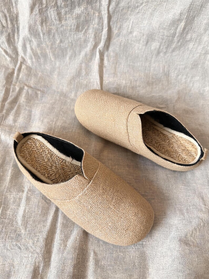 May include: A pair of beige slip-on shoes featuring a textured fabric upper, a rounded toe, and a woven jute insole. The interior lining is black, and the shoes have a low-profile design with a slip-on construction for easy wear and comfort.