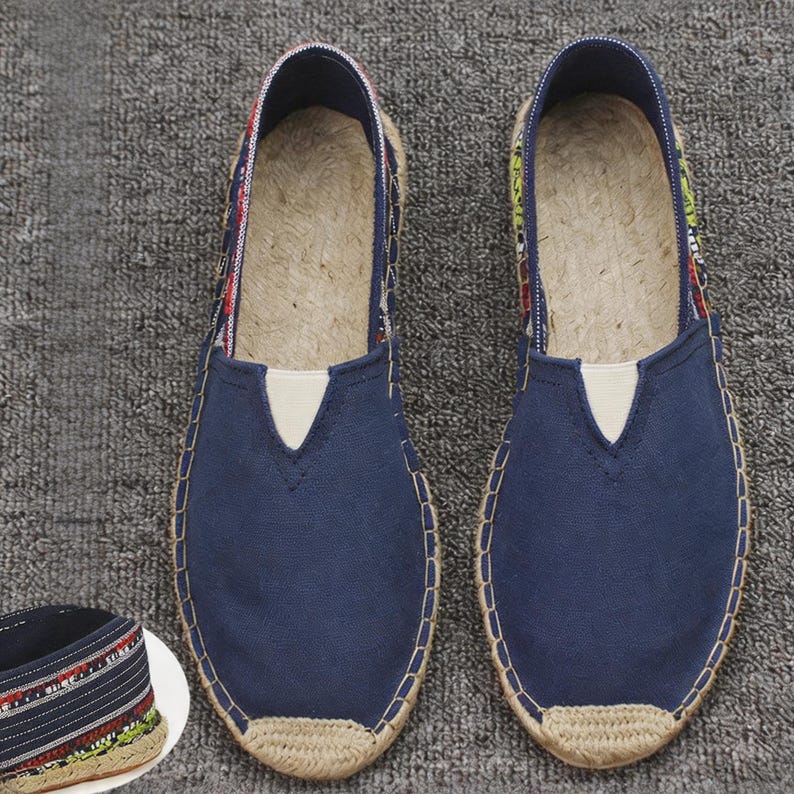 K&ouml;nnte beinhalten: Ein Paar marineblaue Espadrilles aus Segeltuch mit einer Sohle aus geflochtenem Jute und einem beigen elastischen Einsatz am Spann. Das Innenfutter zeigt einen bunten gemusterten Stoff mit roten, gr&uuml;nen und wei&szlig;en Akzenten am Rand.