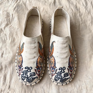 Puede incluir: Un par de zapatos sin cordones de color blanco roto con bordados intrincados de tigres estilizados y patrones abstractos en azul oscuro en la puntera. Los zapatos tienen una parte superior de tela texturizada, punta redonda y suela cosida.
