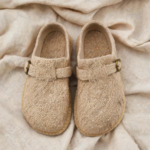 May include: A pair of beige slip-on shoes featuring a textured cable knit fabric upper, a rounded toe, and a decorative strap across the top secured with a small, circular antique brass-toned metal buckle. The shoes have a light-colored stitched trim.