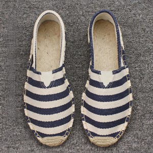 K&ouml;nnte beinhalten: Ein Paar Espadrilles zum Hineinschl&uuml;pfen mit einem horizontalen Streifenmuster in Marineblau und Wei&szlig;. Die Schuhe haben eine abgerundete Zehenpartie, eine Sohlenkante aus geflochtenem Juteseil und ein hellbeiges Innenfutter mit Naturfaser-Fu&szlig;bett.