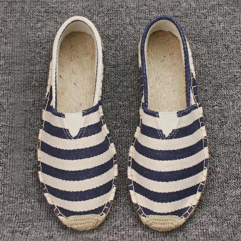 K&ouml;nnte beinhalten: Ein Paar Espadrilles zum Hineinschl&uuml;pfen mit einem horizontalen Streifenmuster in Marineblau und Wei&szlig;. Die Schuhe haben eine abgerundete Zehenpartie, eine Sohlenkante aus geflochtenem Juteseil und ein hellbeiges Innenfutter mit Naturfaser-Fu&szlig;bett.