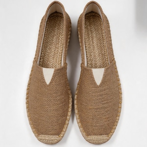 K&ouml;nnte beinhalten: Ein Paar hellbraune Espadrilles mit einer gewebten Jute-Oberfl&auml;che, einer verst&auml;rkten Zehenkappe und einem wei&szlig;en, dreieckigen elastischen Einsatz am Rist. Die Sohlen sind mit geflochtenem Juteseil umwickelt und weisen gelbe Ziern&auml;hte auf.