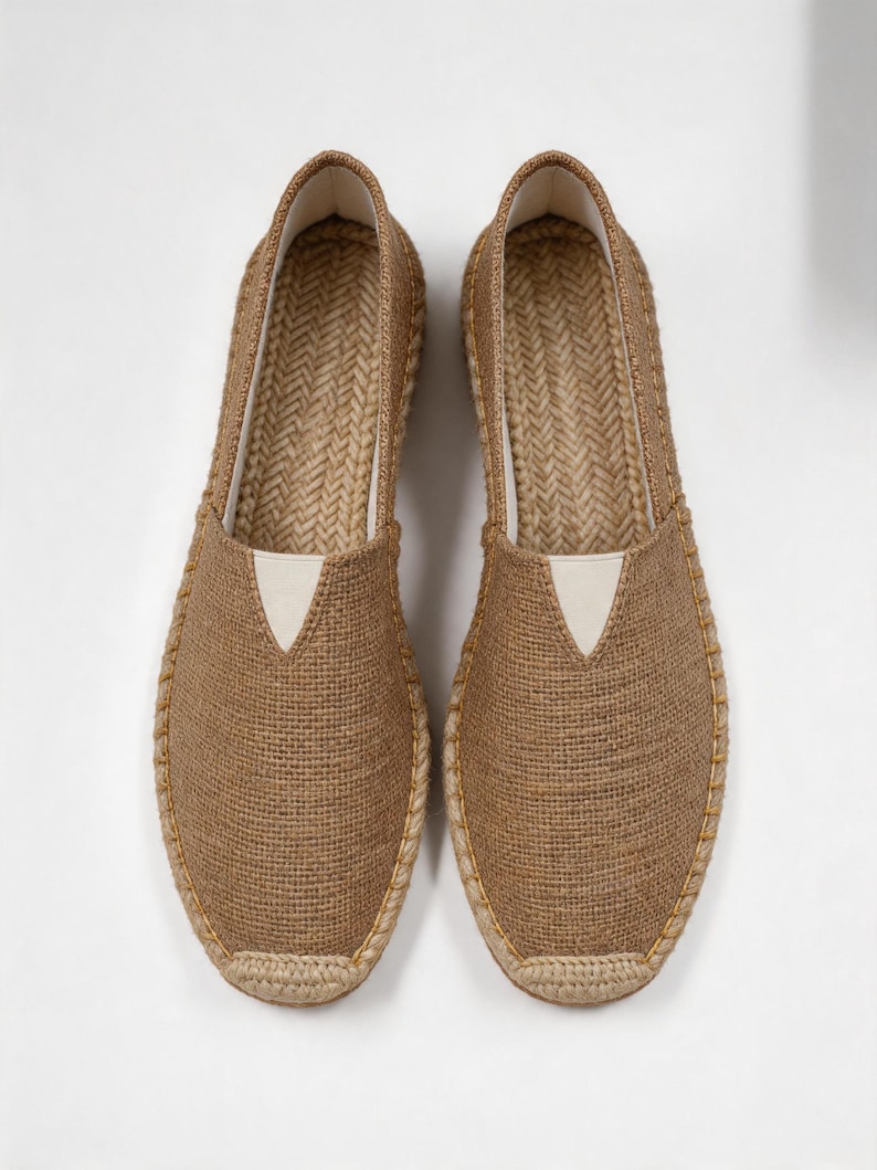 K&ouml;nnte beinhalten: Ein Paar hellbraune Espadrilles mit einer gewebten Jute-Oberfl&auml;che, einer verst&auml;rkten Zehenkappe und einem wei&szlig;en, dreieckigen elastischen Einsatz am Rist. Die Sohlen sind mit geflochtenem Juteseil umwickelt und weisen gelbe Ziern&auml;hte auf.