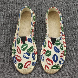 K&ouml;nnte beinhalten: Ein Paar Slipper-Espadrilles mit einem cremefarbenen Canvas-Obermaterial und einem sich wiederholenden Muster aus roten, gr&uuml;nen und blauen Lippenabdr&uuml;cken. Die Schuhe haben eine Sohle aus geflochtener Jute, eine runde Spitze und einen schwarzen Gummizug.