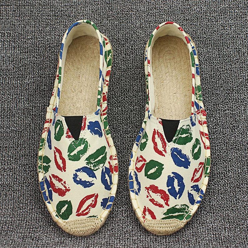 K&ouml;nnte beinhalten: Ein Paar Slipper-Espadrilles mit einem cremefarbenen Canvas-Obermaterial und einem sich wiederholenden Muster aus roten, gr&uuml;nen und blauen Lippenabdr&uuml;cken. Die Schuhe haben eine Sohle aus geflochtener Jute, eine runde Spitze und einen schwarzen Gummizug.
