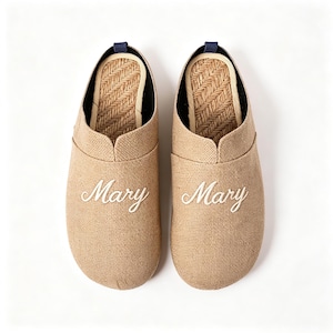 May include: A pair of beige slip-on slippers with a textured fabric exterior and a woven herringbone pattern on the inner sole. Each slipper features the name Mary embroidered in white cursive script on the front and a small dark blue heel tab.