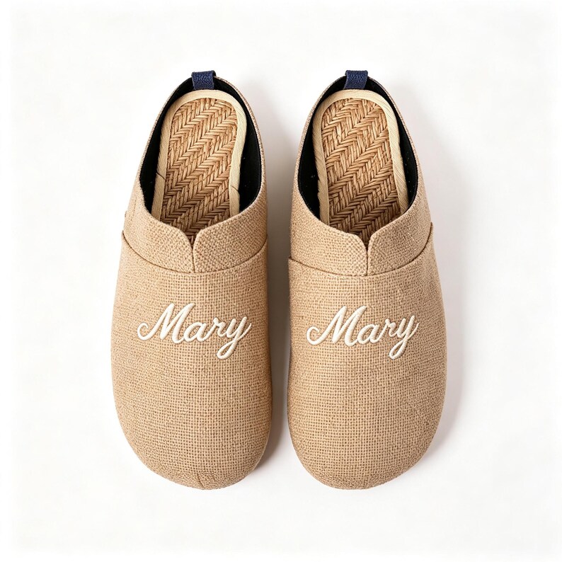 May include: A pair of beige slip-on slippers with a textured fabric exterior and a woven herringbone pattern on the inner sole. Each slipper features the name Mary embroidered in white cursive script on the front and a small dark blue heel tab.