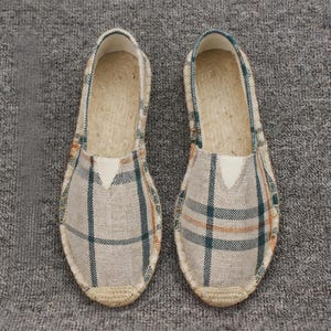 K&ouml;nnte beinhalten: Ein Paar Slipper mit beigem Stoffobermaterial und einem dunkelgr&uuml;nen sowie orangefarbenen Karomuster. Die Schuhe verf&uuml;gen &uuml;ber eine geflochtene Jute-Umrandung an der Sohle und einen hellen, dreieckigen elastischen Einsatz am Vorderblatt.