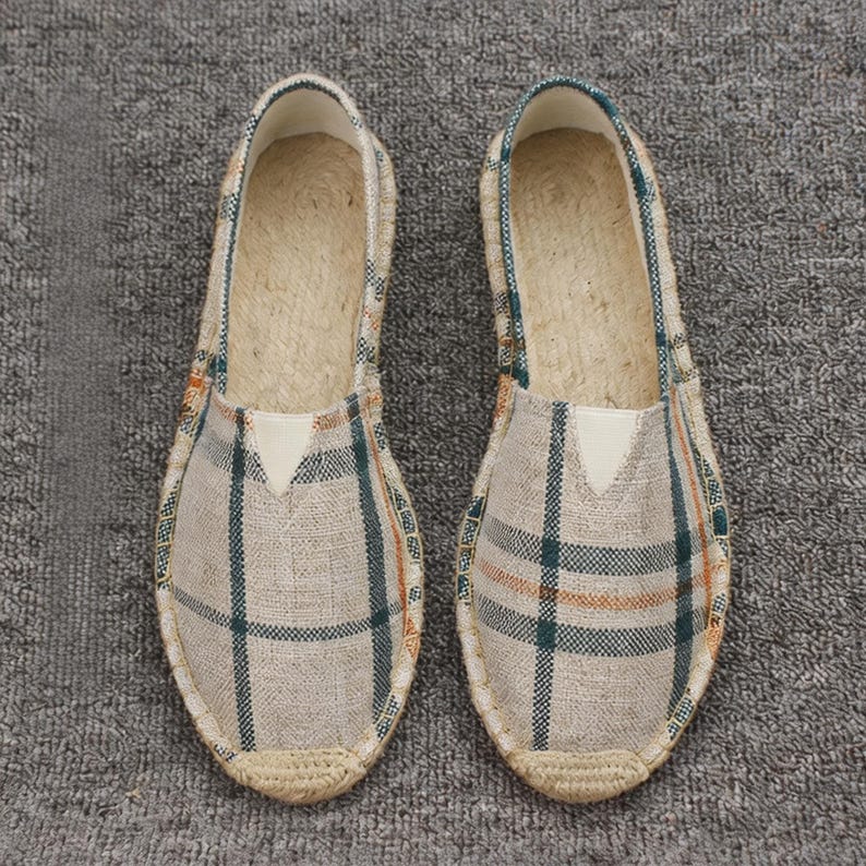 K&ouml;nnte beinhalten: Ein Paar Slipper mit beigem Stoffobermaterial und einem dunkelgr&uuml;nen sowie orangefarbenen Karomuster. Die Schuhe verf&uuml;gen &uuml;ber eine geflochtene Jute-Umrandung an der Sohle und einen hellen, dreieckigen elastischen Einsatz am Vorderblatt.