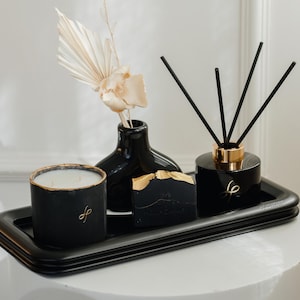 May include: A black tray displays a black candle with gold edging, a black vase with dried flowers, a black soap bar with gold detailing, and a black diffuser with reeds. The items are on a white surface.