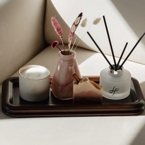May include: A wooden tray holds a candle, a small vase with dried flowers, a bar of soap, and a diffuser. The candle is white with a gold rim. The vase is pink. The soap is brown and tan. The diffuser is frosted glass.