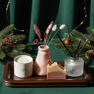 Op de afbeelding: Een donkerhouten dienblad toont een witte kaars met een gouden rand, een roze vaas met gedroogde bloemen, een stuk zeep en een diffuser van matglas met zwarte stokjes. De items staan op een groene ondergrond met kerstversiering.