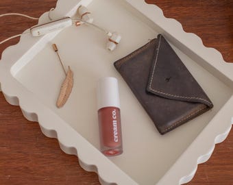 May include: A white, scalloped-edge tray contains a brown leather wallet, a lipstick tube with "cream co." printed on it, a gold feather, and white earbuds. A frosted glass diffuser is visible in the background.